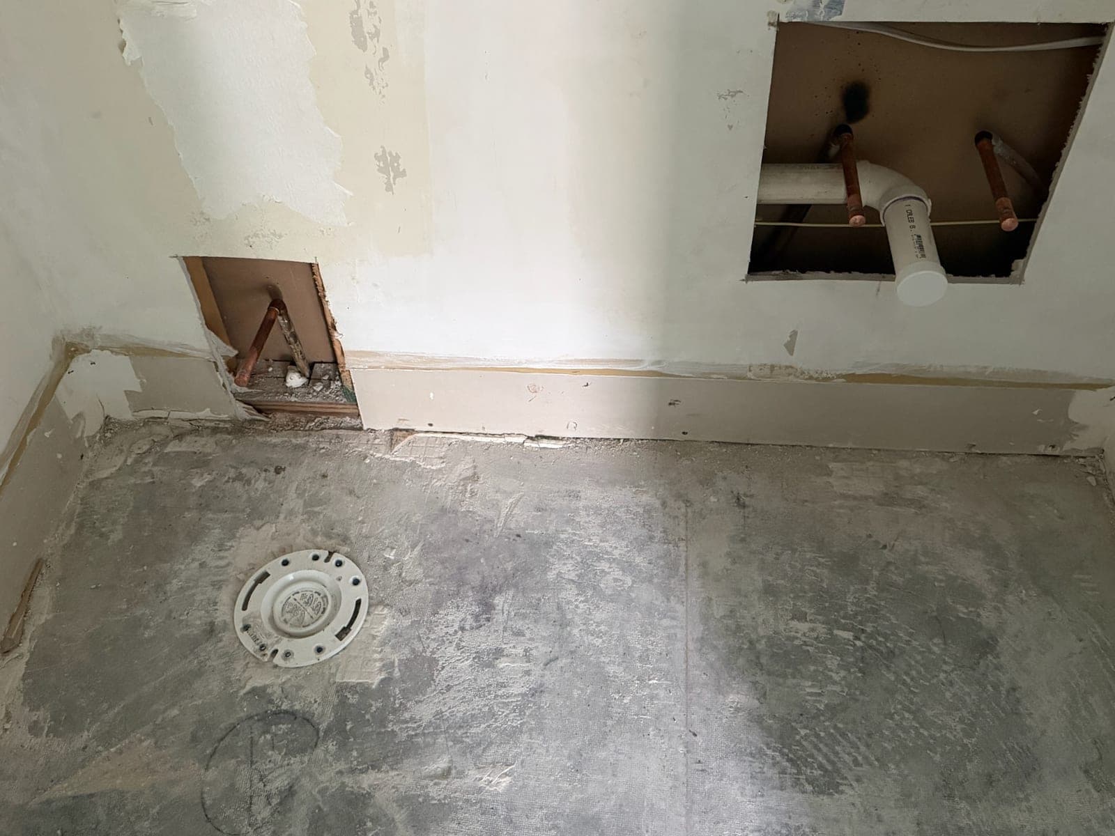 Toilet closet flange set into a bare concrete slab, with wall access cuts exposing copper supply and a capped PVC waste line