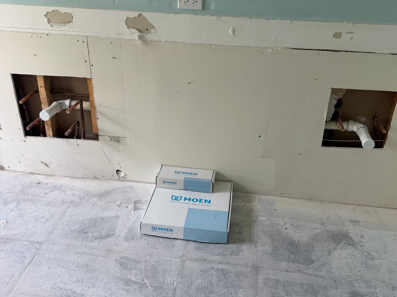 Two neat access openings cut in a wall exposing copper supply and PVC waste lines, with new fixtures boxed on the floor